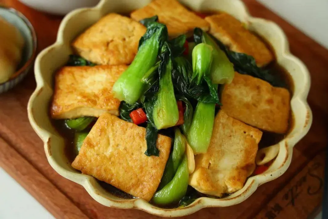 Fried tofu pieces and bok choy in a dark sauce served in a scalloped bowl, the colour of the sauce the only thing on the table that cannot be identified by eye