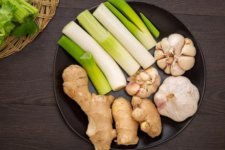 A black plate laid out with whole spring onions, garlic bulbs, a full head of garlic and ginger roots, with celery and leafy greens in a basket behind