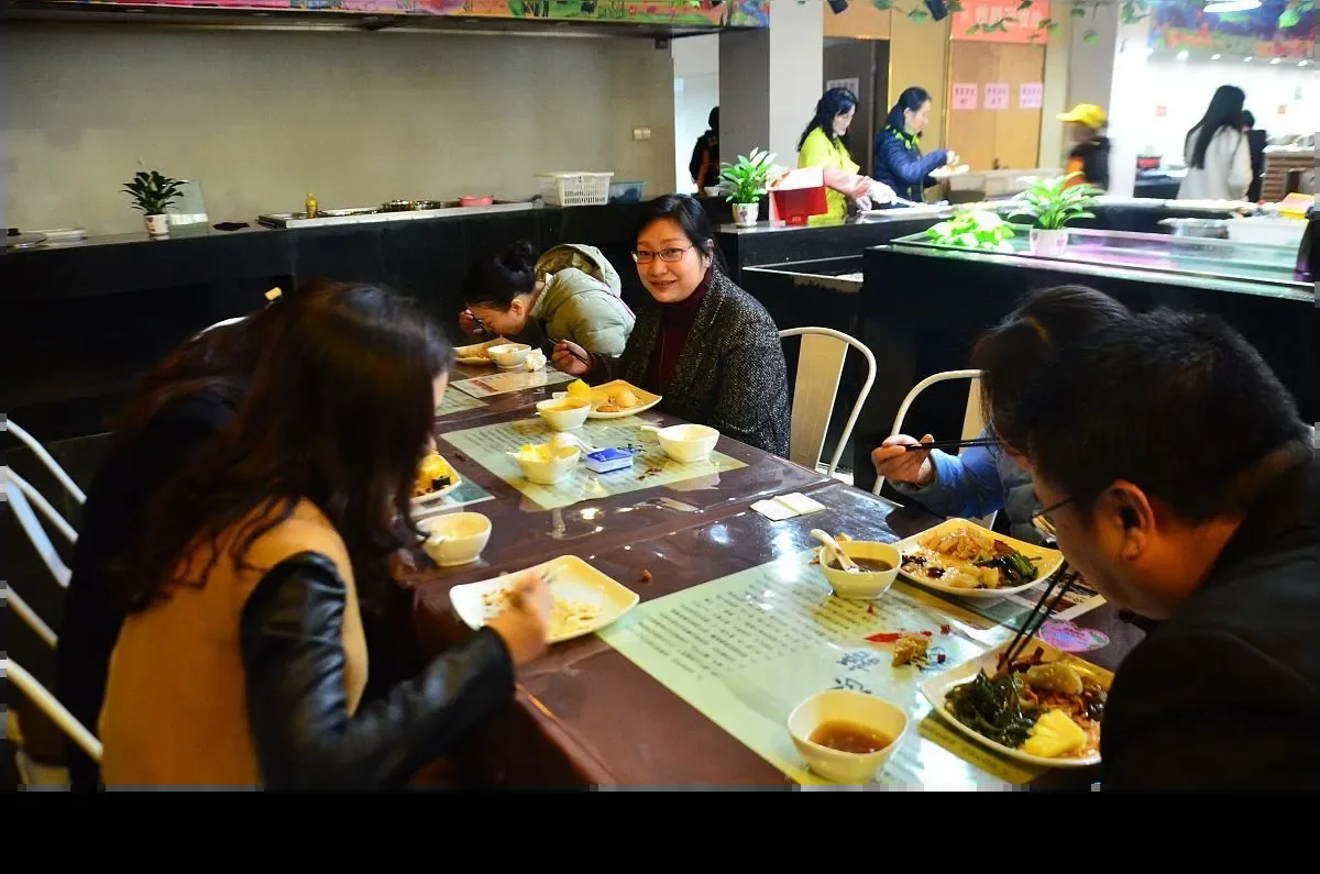 A busy self-service restaurant with several groups eating rice and shared vegetable dishes at long tables, and a serving counter with staff along the back