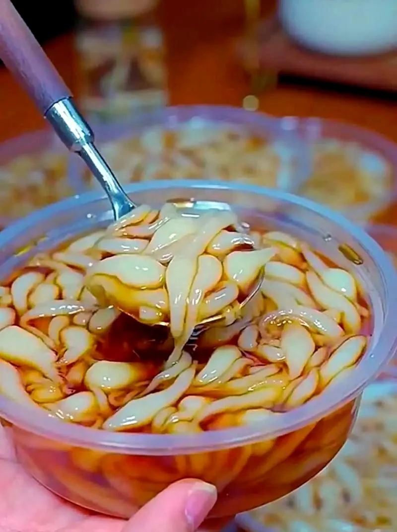 White rice jelly pieces shaped like small shrimp floating in brown sugar water in a clear plastic cup, lifted on a spoon