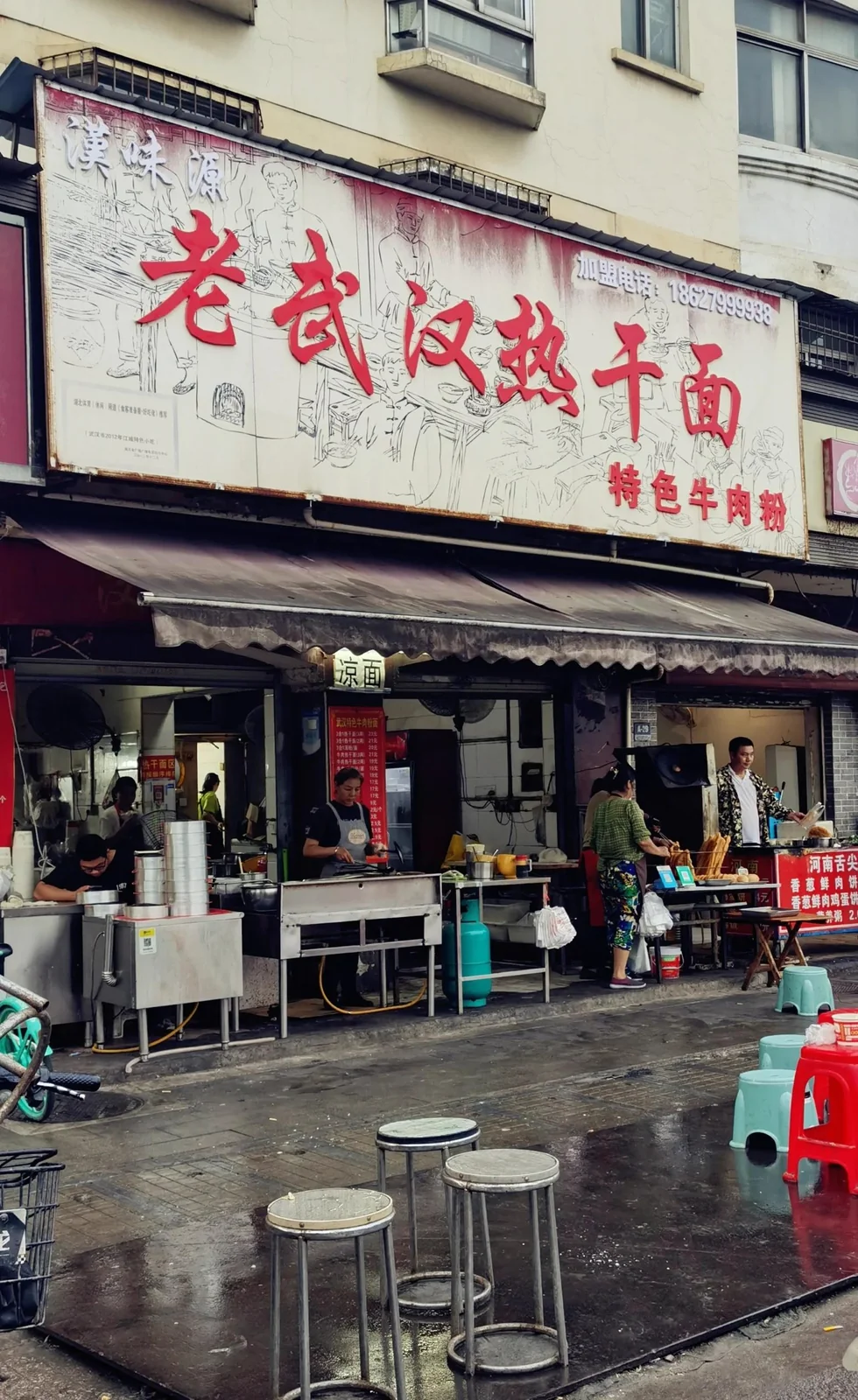 A street-side noodle shop under a large painted sign, with staff working at counters and plastic stools on wet pavement