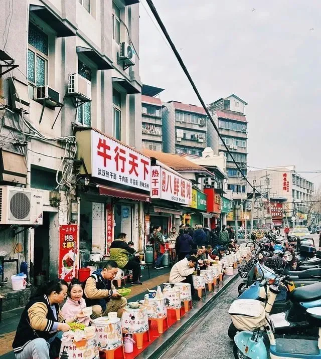 Narrow Wuhan street lined with breakfast stalls where customers eat at low plastic stools beside parked scooters