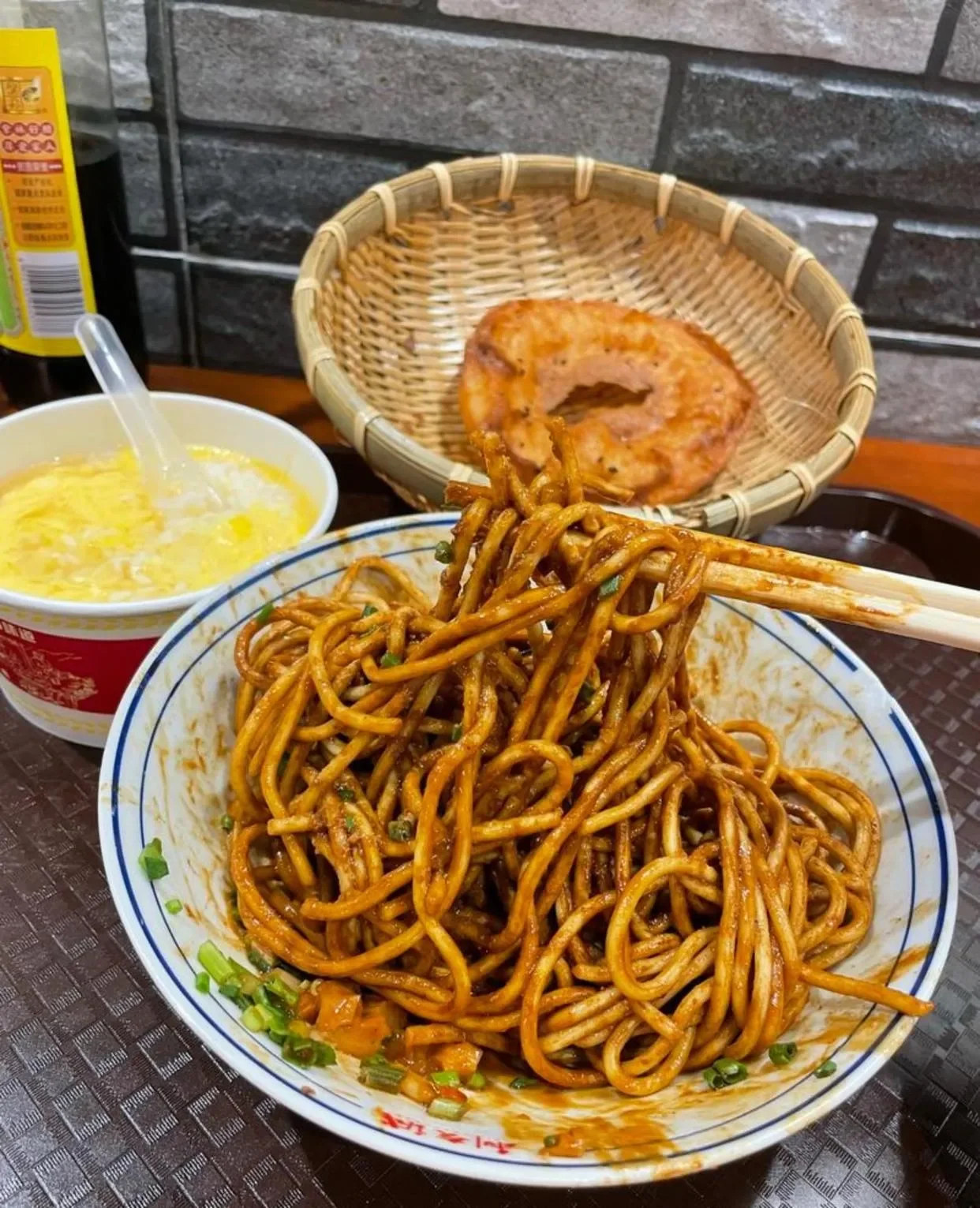 Bowl of sesame-coated hot dry noodles lifted with chopsticks beside a paper cup of egg drop soup and two fried mianwo in a bamboo basket