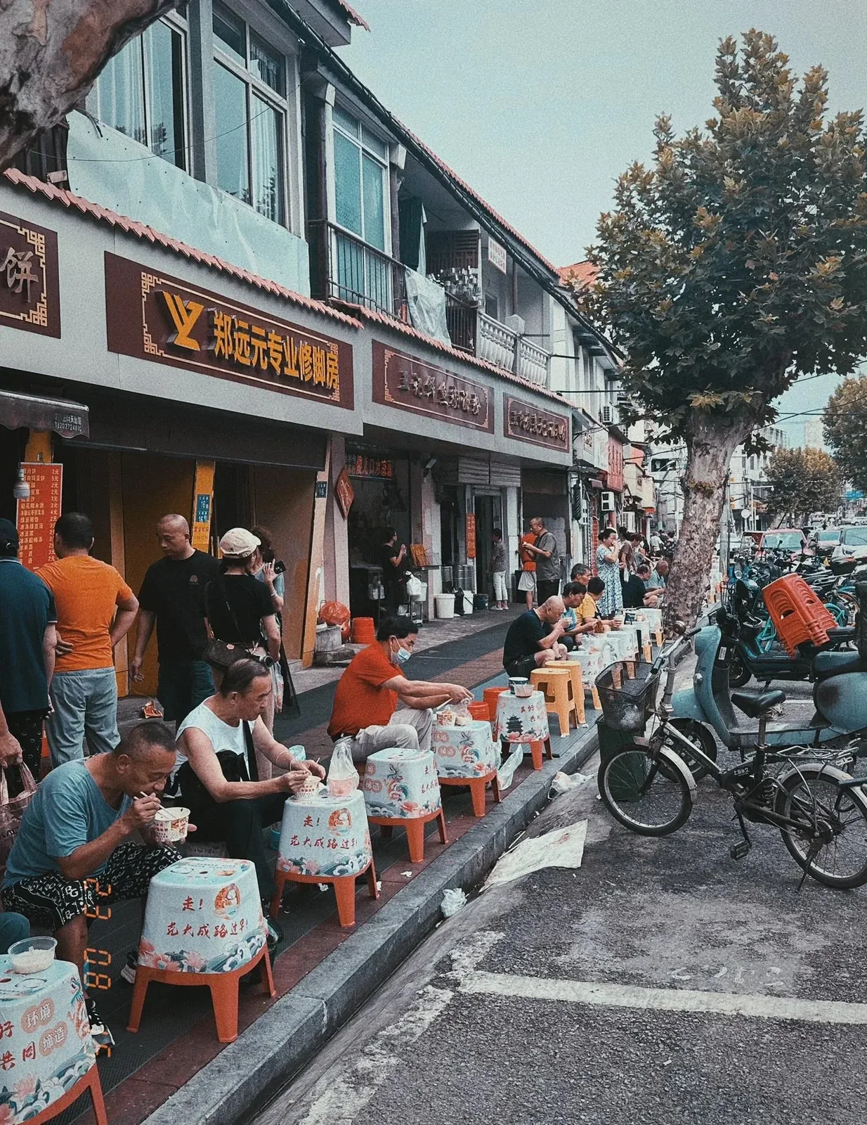Customers eating at low vinyl-covered plastic stools along a Wuhan sidewalk beside parked bicycles and scooters