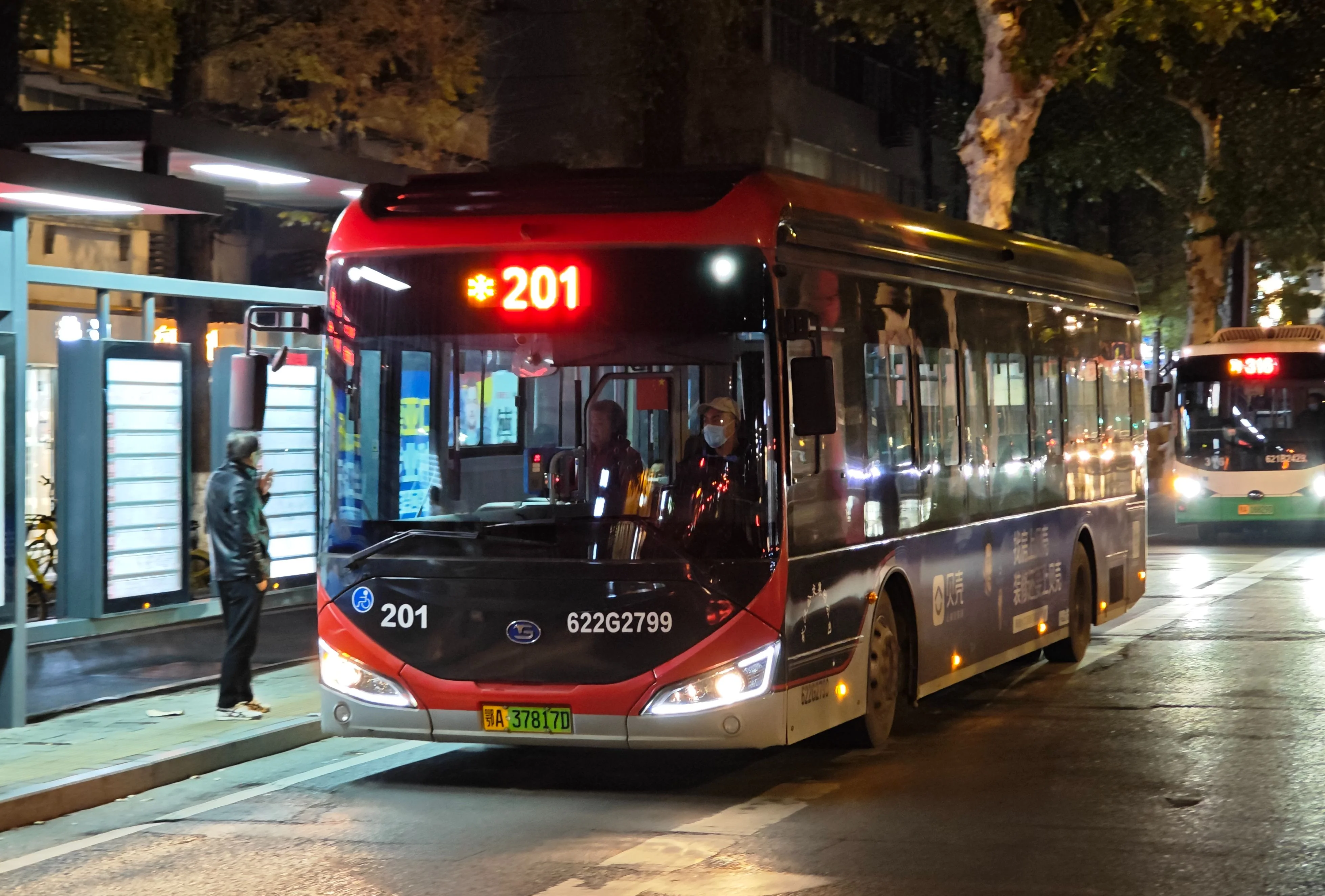 A Wuhan bus at night driving down a brightly lit boulevard, its windows glowing, motion blur suggesting speed, neon signs reflecting on wet pavement