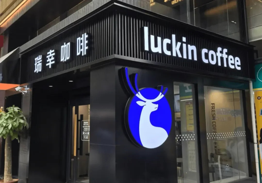 A Luckin Coffee storefront on a city street, with the blue deer logo lit up and the name in both English and Chinese on a black facade