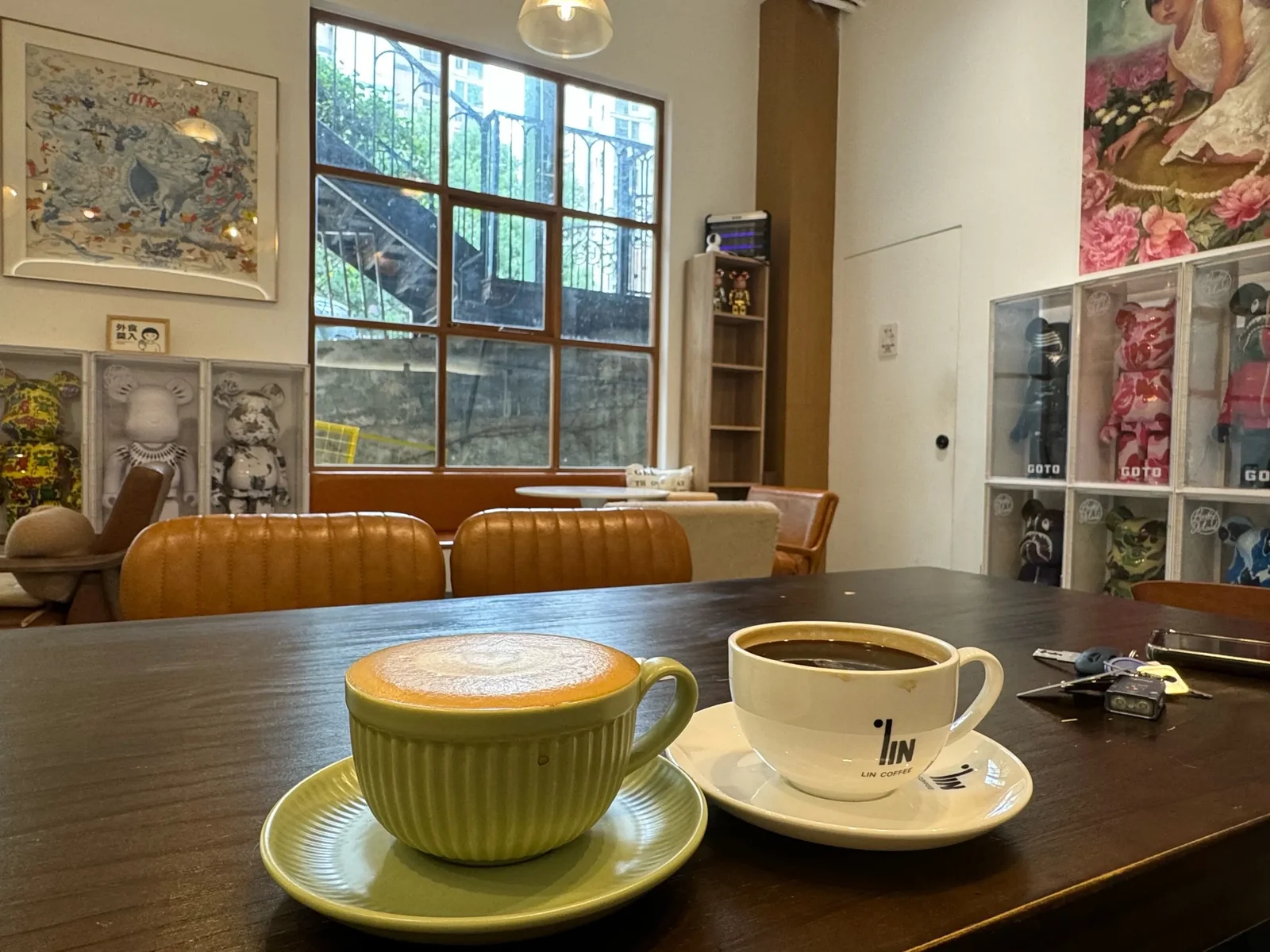 Inside an independent cafe, a ceramic cup of coffee and a white paper cup on a dark wooden table in front of a tan leather bench, with framed art and shelves of designer figures behind