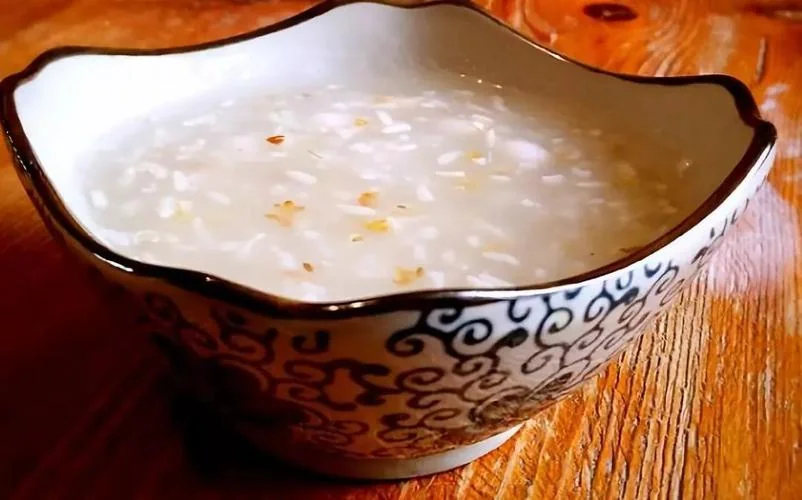 A bowl of glutinous rice paste wine in a blue-and-white porcelain bowl on a wooden table, the rice grains visible in a thick milky base with small golden flecks scattered through it
