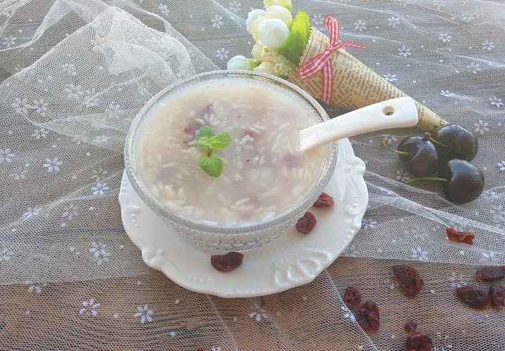 A bowl of glutinous rice soup topped with goji berries and a mint sprig on an ornate white saucer with a ceramic spoon, staged on a lace cloth