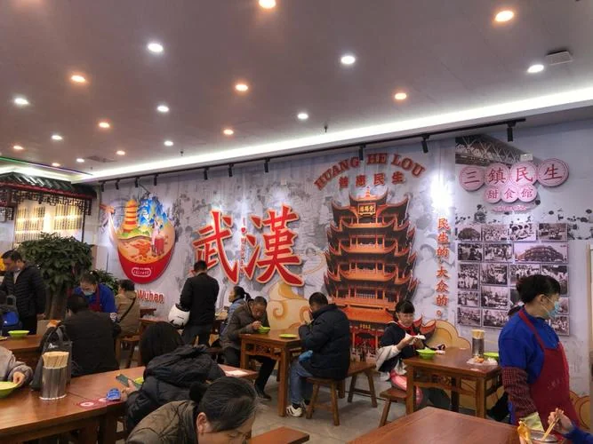 The interior of a busy Wuhan sweet shop, customers seated at wooden tables eating from green bowls, staff in blue uniforms and red aprons, and a wall mural reading 三镇民生甜食馆 beneath a Yellow Crane Tower illustration