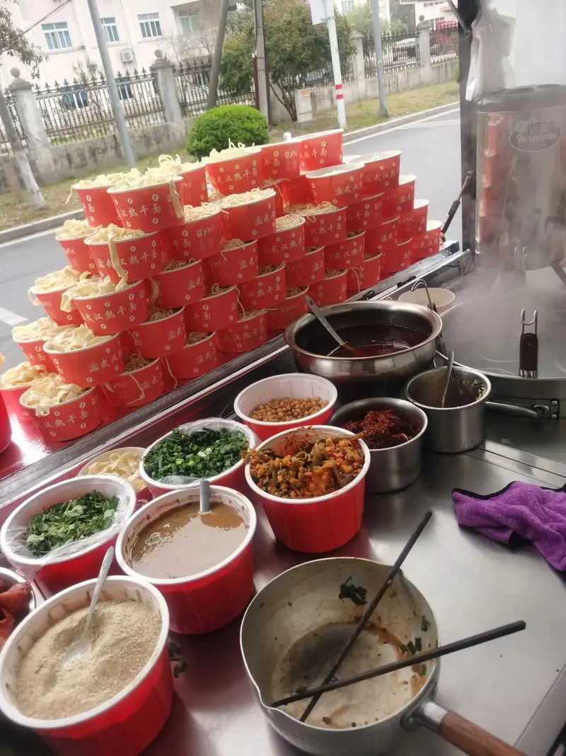 A Wuhan noodle stall auntie serving hot dry noodles while calling out to customers, capturing the loud and lively atmosphere of a Wuhan breakfast stall