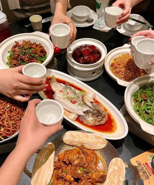 A crowded Wuhan restaurant table with baijiu bottles, small glasses, and people mid-toast, hands raised, faces animated