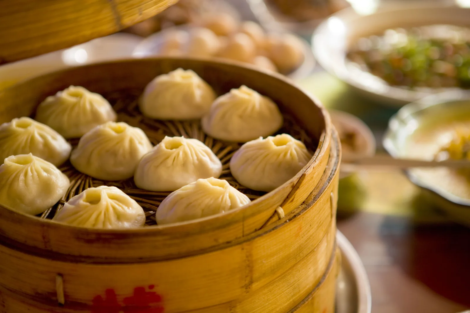 Nine plump white soup dumplings with pleated tops sitting in a round bamboo steamer under warm low light, steam rising, with a second basket and small condiment dishes blurred behind