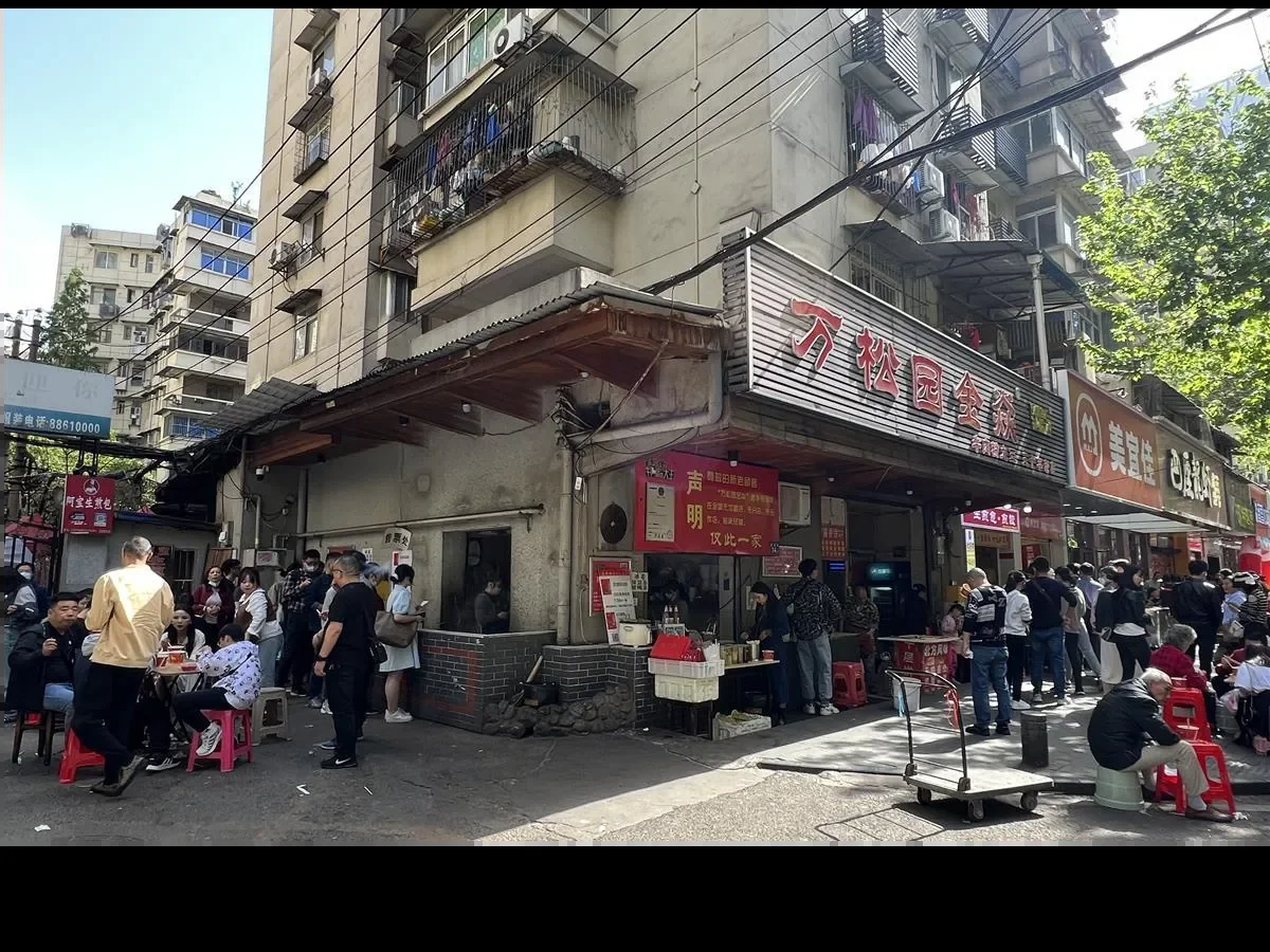 The Wansongyuan Food Street sign above a crowded pavement breakfast scene, customers seated on red plastic stools beside a serving cart, with residential balconies and power lines overhead