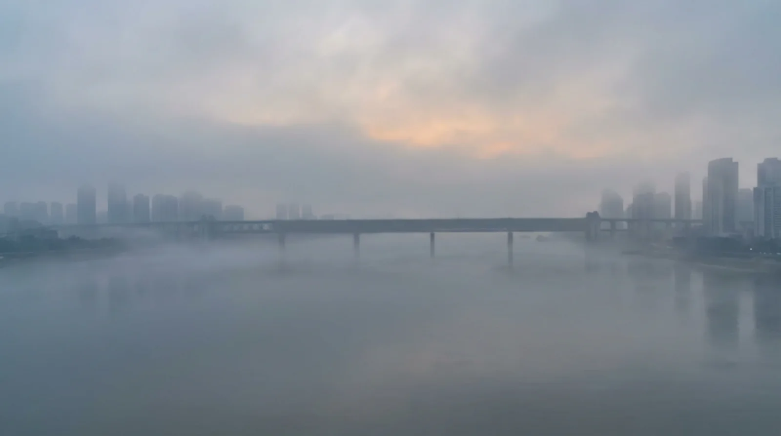 Winter fog scene of a wide river with a bridge crossing through mist, distant buildings barely visible as silhouettes