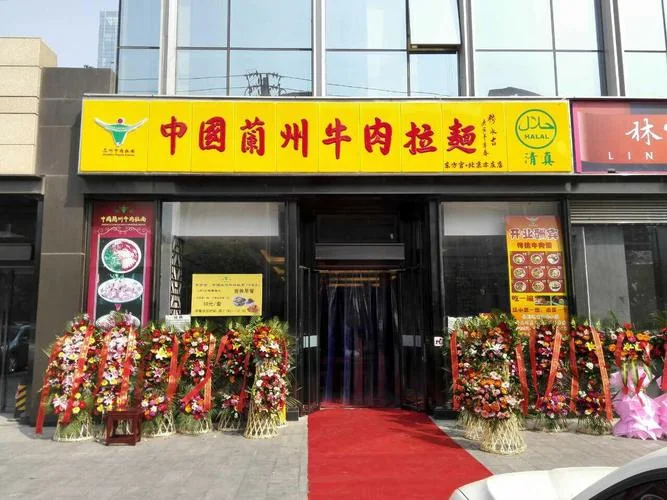 A Lanzhou beef noodle shopfront with a yellow signboard, red lettering, and a green 清真 halal mark with the Arabic halal symbol, flanked by congratulatory flower baskets and a red carpet