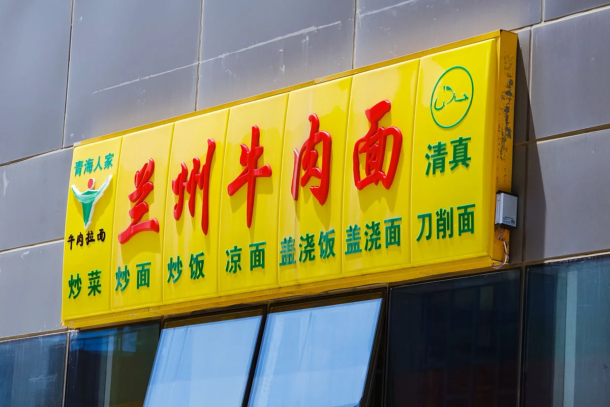 A close-up of a yellow signboard reading 兰州牛肉面 in large red characters with a green 清真 mark and the Arabic halal symbol in a circle, above a menu of stir-fried dishes, cold noodles and rice