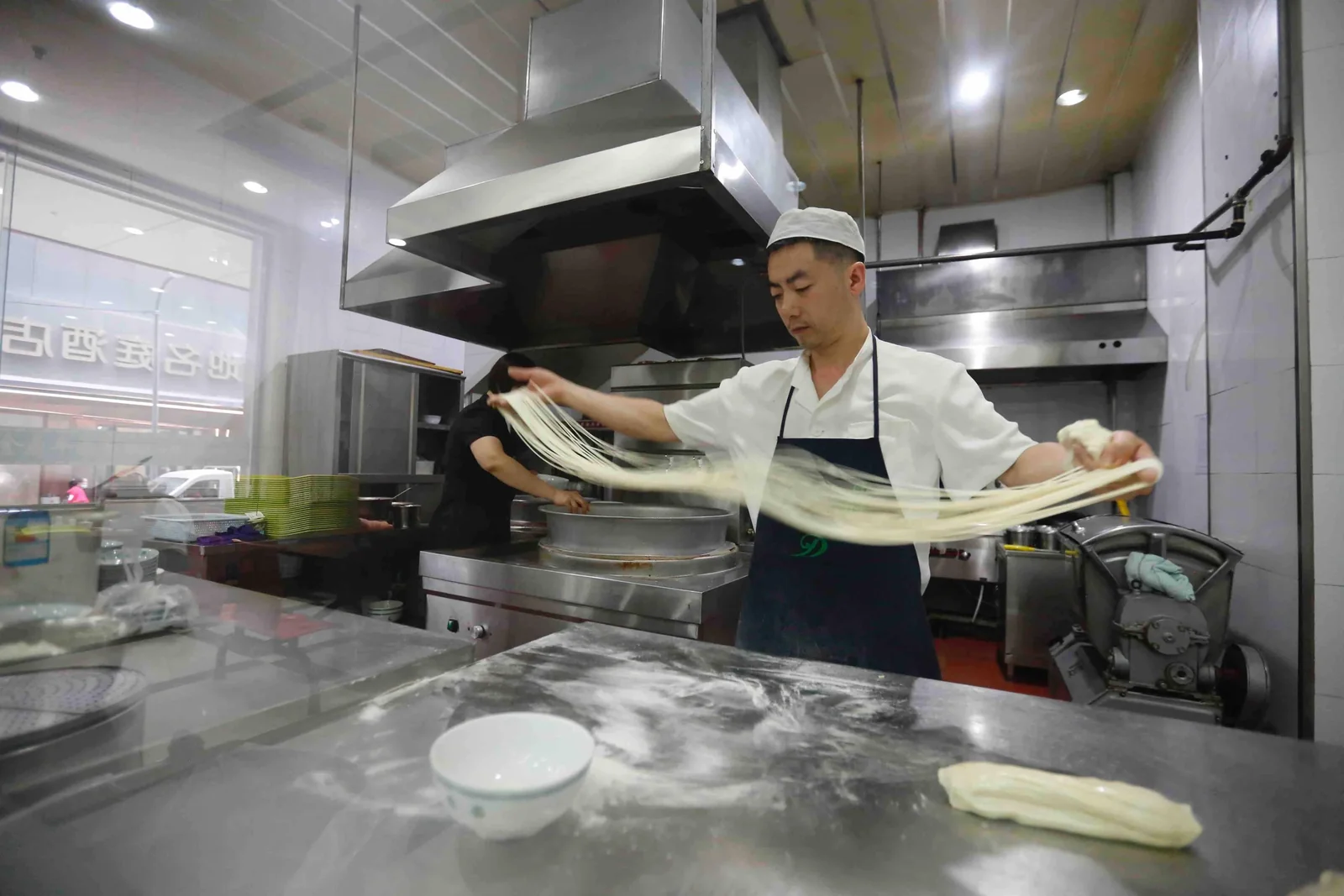 A cook in a white cap and apron stretching a long piece of dough by hand over a floured steel counter in an open restaurant kitchen, with extraction hoods and stacked bowls behind him