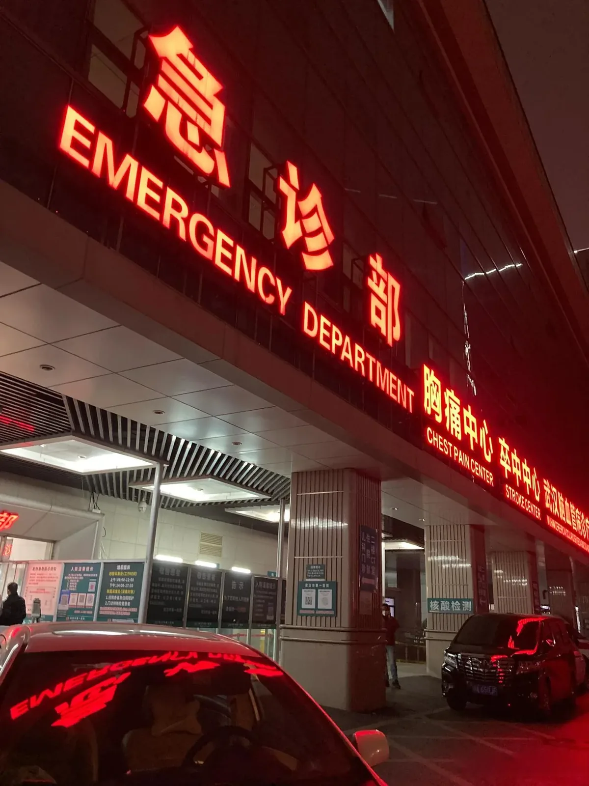 The emergency entrance of a Wuhan hospital at night, lit up and active, where patients and staff move through the 24-hour urgent care system