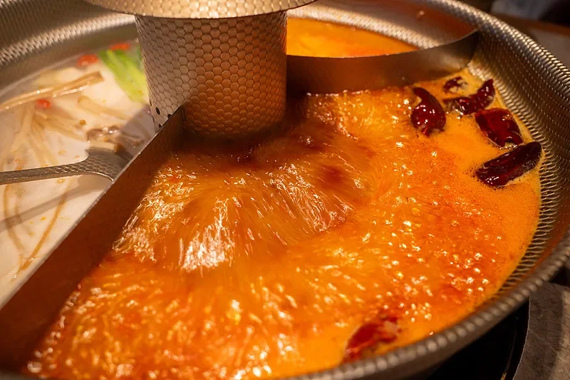 A divided hot pot seen from above, with a clear broth on one side and an orange-red chilli oil broth on the other, separated by a metal chimney-style divider with a mesh skimmer resting across it