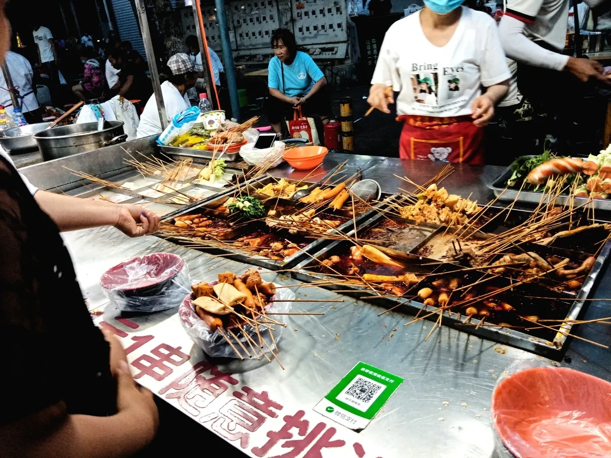 A street stall laid out with rows of bamboo skewers holding grilled meat and vegetables, with customers selecting skewers at the counter and a vendor in an apron behind it