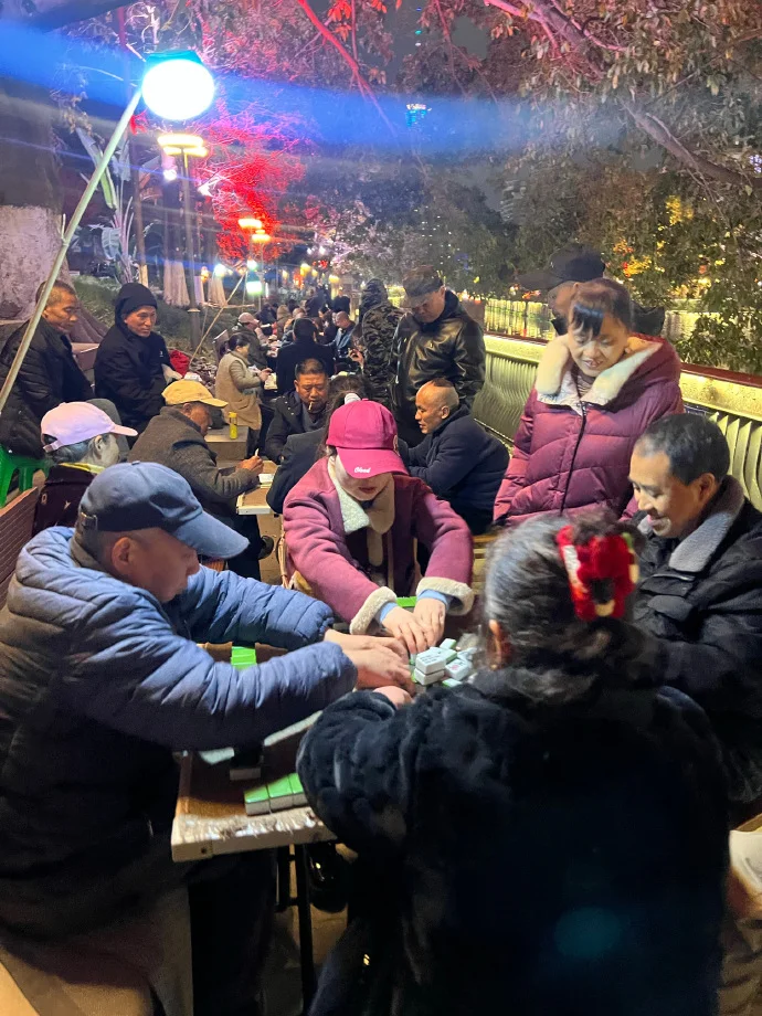 People playing mahjong at a street-side table under an awning in Wuhan, with onlookers and a nearby barbecue grill