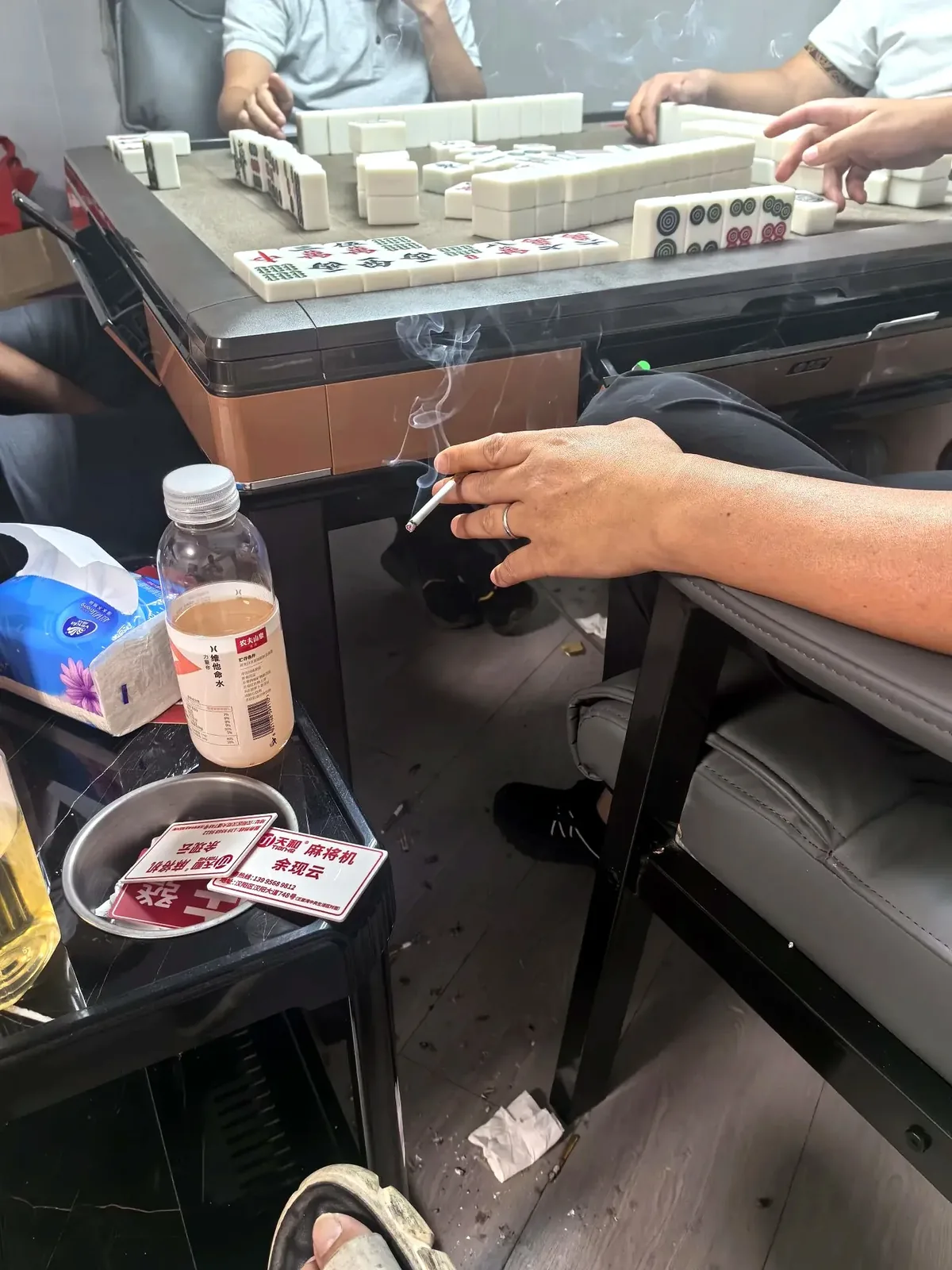 Close-up of table corner showing paper scorecard, 5 RMB and 10 RMB notes, cigarette pack, and a half-empty tea cup