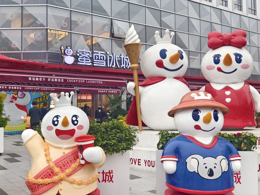 Mixue milk tea shop on a busy Chinese street, showing the iconic snowman mascot signage and a line of customers, the budget tea chain that dominates with 4-yuan drinks