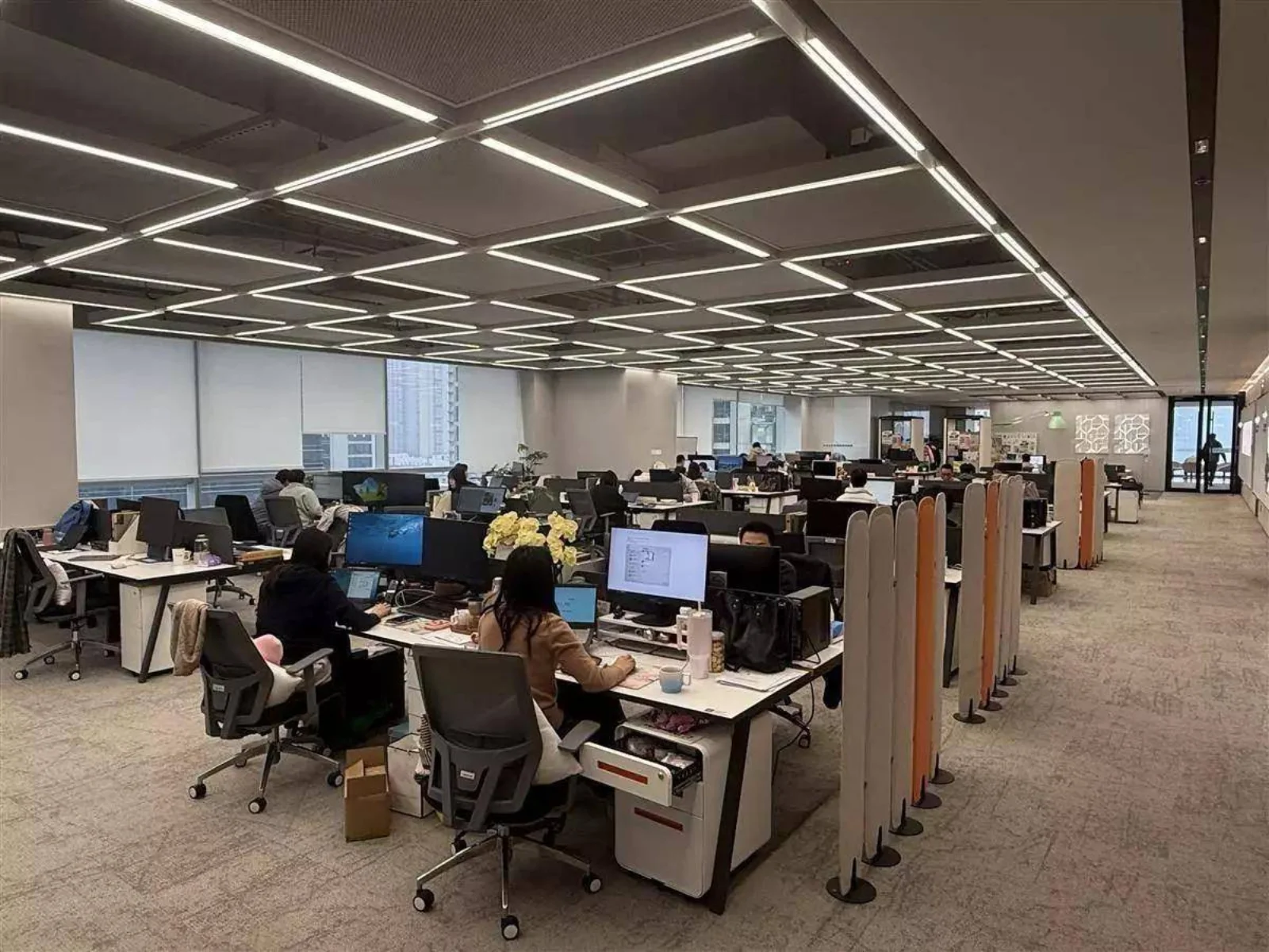 Modern open-plan office space with long rows of white desks, employees working at monitors and laptops, grid-pattern LED ceiling lights above