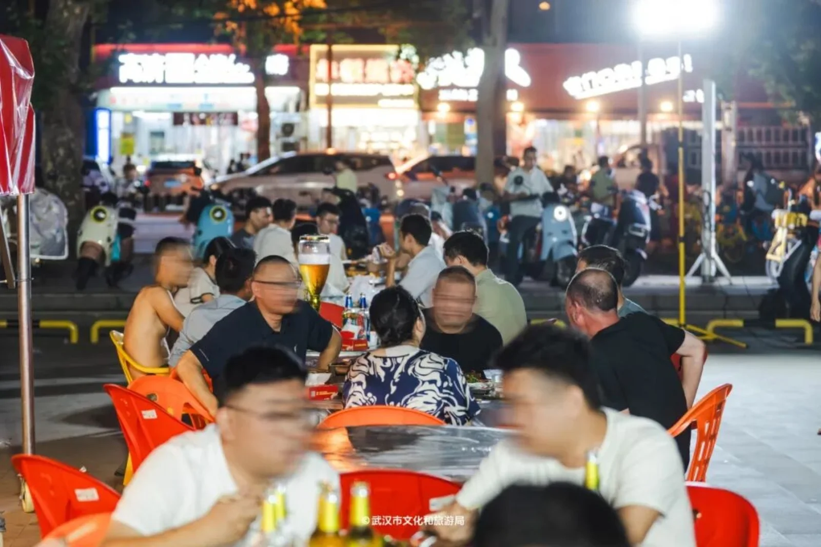 Two older men sitting at a small Wuhan street table with beer bottles, one gesturing emphatically mid-argument, both laughing despite the animated conversation