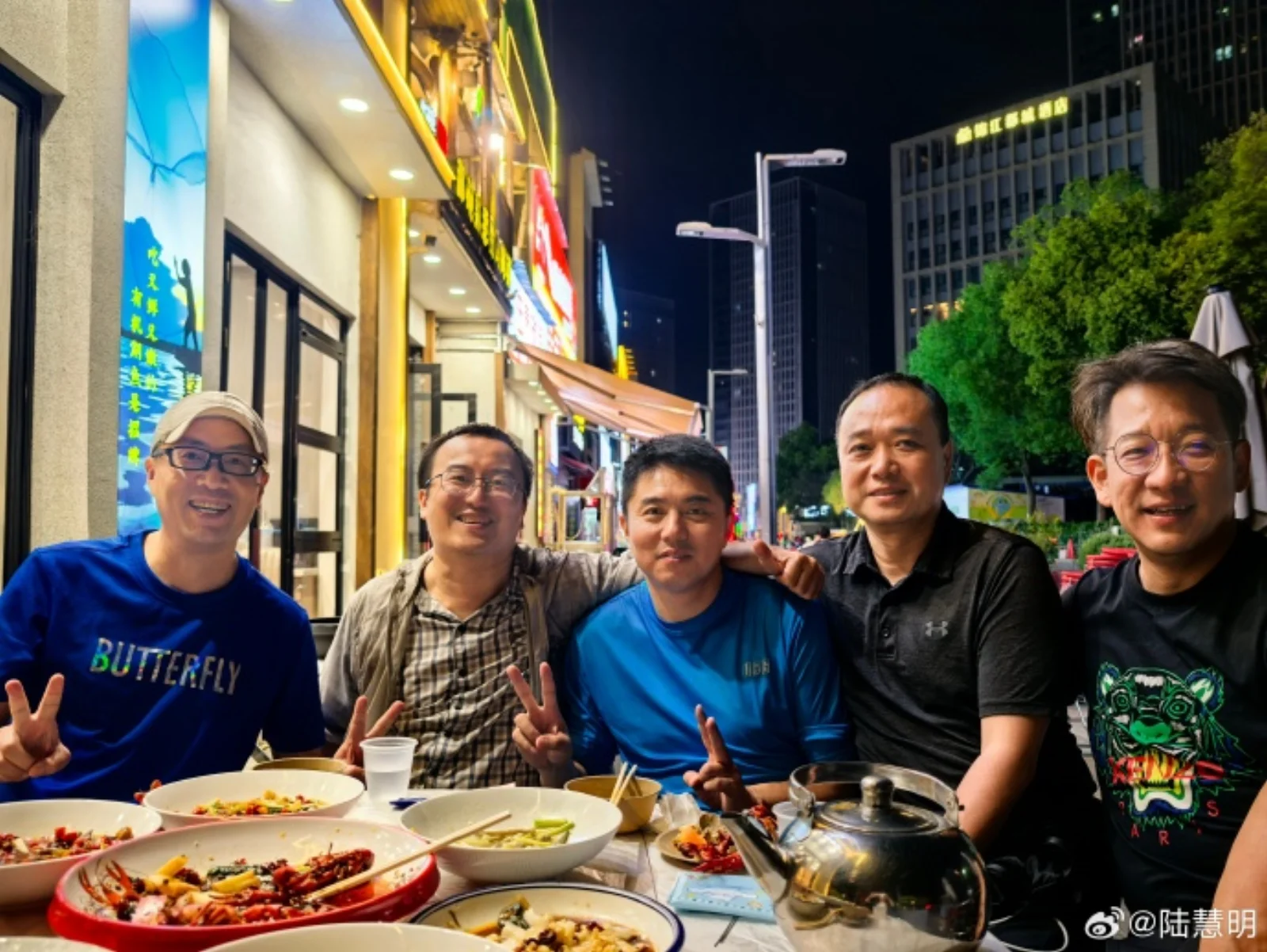 A group of friends gathered around a late-night Wuhan food table, faces lit by warm yellow light, raising beer cans in a toast, laughing with mouths wide open