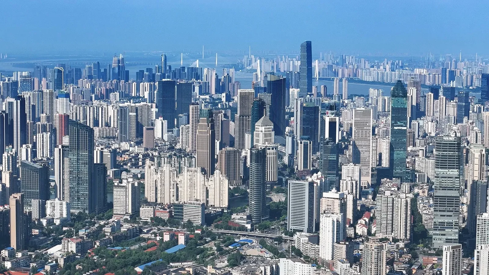 The Wuchang skyline seen from above in haze, the 475-metre Wuhan Greenland Center rising right of centre, with the Yangtze behind it, two cable-stayed bridges with white A-frame pylons, and dense residential towers receding into the distance