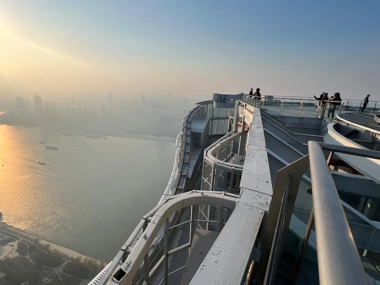 Visitors at a curved steel railing on a high observation deck looking down over a wide river with boats, hazy skyline beyond, and low sun flaring on the left