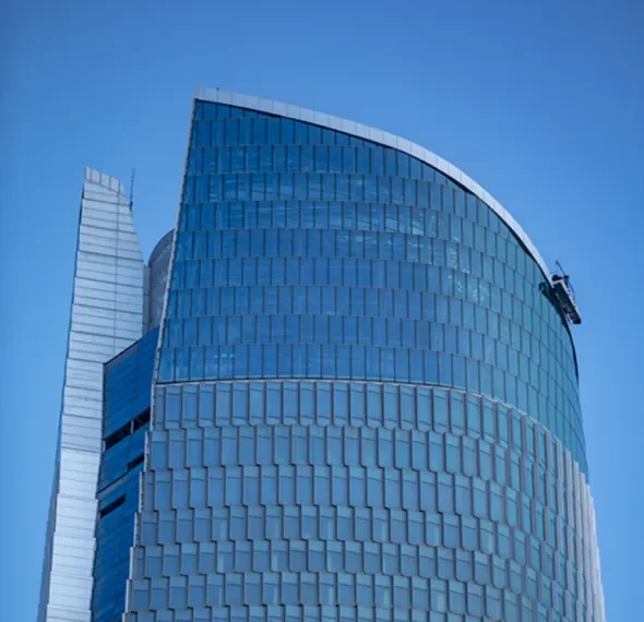 Looking up at a curved blue glass office tower with a white capped crown against clear blue sky, with a slimmer white ribbed tower rising behind it