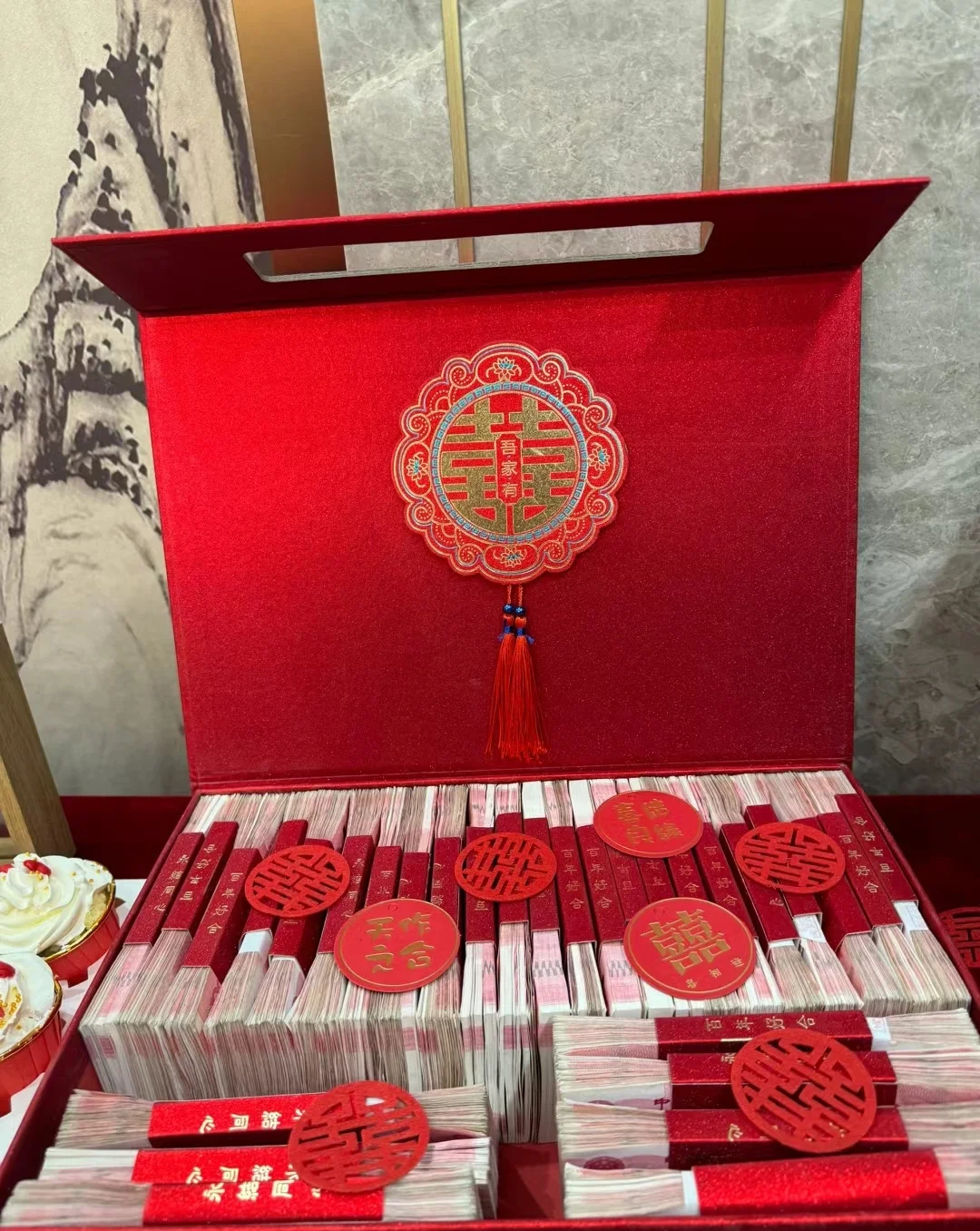 Red envelopes with cash gifts being collected or piled on a table next to a guest registry, with hosts in formal attire smiling and toasting, showing the critical red envelope economy that funds and measures social reciprocity at a Wuhan celebration