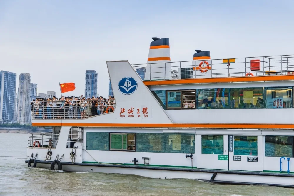 A white and green passenger ferry crowded with people along the upper deck railing, a Chinese flag at the stern and apartment towers on the far bank